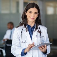 Portrait of beautiful mature woman doctor holding digital tablet and looking at camera. Confident female doctor using digital tablet with colleague talking in background at hospital. Latin nurse in labcoat and stethoscope in private clinic with medical team working.